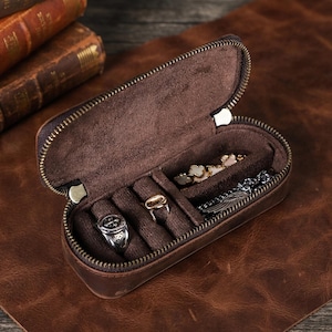 May include: An open, oval-shaped brown leather jewelry case with a zipper closure. The interior is lined with dark brown fabric and features dedicated slots for rings and a small compartment containing several silver-toned rings and a metal chain.
