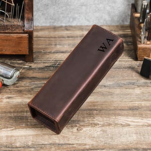 May include: A brown leather case with the initials "WA" embossed on the front. The rectangular case is designed to hold a long, slender object. The leather has a distressed look, and the case is sitting on a wooden surface.