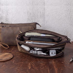 May include: A brown leather zipped pouch with multiple interior compartments containing a stack of paper currency, a smartphone, a white charging block, a coiled white charging cable, and a white wireless earbud case, with a matching pouch behind.