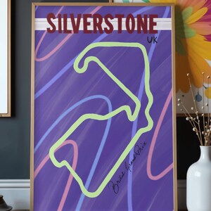 May include: A framed poster featuring the Silverstone race track layout in neon green on a purple background. The word "SILVERSTONE" is in maroon, with "UK" below. Pink and blue lines swirl across the background. The words "British Grand Prix" are handwritten.