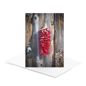 May include: A vibrant red chili ristra, dusted with snow, hangs against a weathered wooden wall. The ristra is tied with natural twine, showcasing the rustic charm of Southwestern decor. The image is a greeting card.
