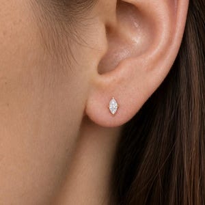 May include: A small marquise-cut diamond stud earring set in a thin gold metal post. The earring is positioned in the lower earlobe, featuring a single clear, faceted stone with pointed ends that align vertically along the center of the lobe.