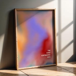 May include: A rectangular framed print featuring a vibrant, blurred gradient of purple, orange, and red colors. The text "THE EVER EXPANDING NOW" is printed in white, sans-serif capital letters in the lower right corner of the artwork.