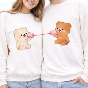 May include: White sweatshirts featuring a cartoon bear design. One bear is light beige, the other is brown, each holding a pink cup connected by a red string. The bears appear to be communicating. A cute and playful design.