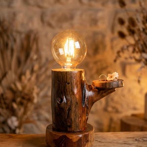 Puede incluir: Lámpara rústica de madera con una bombilla cálida y brillante. La lámpara está hecha de un tronco de árbol, con una pequeña repisa que sostiene dos anillos y un cristal. La lámpara está sobre una superficie de madera.