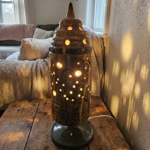 Puede incluir: Una lámpara de mesa de madera con cuerpo cilíndrico y parte superior escalonada. La lámpara tiene diseños florales y circulares tallados con agujeros recortados que emiten un brillo cálido. La lámpara se asienta sobre una base gris oscuro y está enchufada a un cable eléctrico blanco.