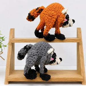 May include: Two crocheted animal figures standing on a two-tiered wooden shelf. The top figure is orange with black accents, and the bottom figure is gray with black accents. Both have black eyes, white facial markings, and striped tails.