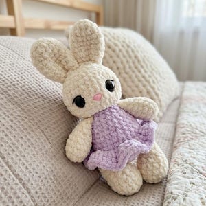 Könnte beinhalten: Ein handgefertigtes Häschen-Plüschtier aus Häkelarbeit. Das Häschen ist hauptsächlich cremefarben mit einem lavendelfarbenen Kleid und schwarzen Augen. Das Häschen sitzt auf einem Bett mit einer gemusterten Decke.
