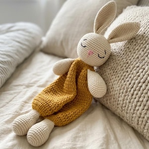 Könnte beinhalten: Ein handgefertigtes Häschen-Spielzeug aus Häkelarbeit in Creme und Senfgelb. Das Häschen hat lange Ohren, geschlossene Augen und eine rosa Nase. Es liegt auf einem Bett mit Kissen. Das Spielzeug ist etwa 30 cm lang.