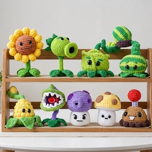 Pode incluir: Uma coleção de nove figuras de pelúcia em crochê inspiradas em personagens de plantas, dispostas em uma prateleira de madeira de dois níveis. As figuras incluem um girassol, um lançador de ervilhas verde, uma planta folhosa, um melão, uma espiga de milho, uma dioneia, um cogumelo roxo, um cogumelo bege e um personagem bulboso marrom.