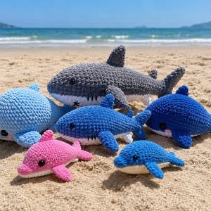 Könnte beinhalten: Eine Sammlung von sechs gehäkelten Plüschtieren in Form von Haien und Delfinen in verschiedenen Farben, darunter Grau, Hellblau, Dunkelblau und Pink. Jedes Stofftier hat eine strukturierte Garnoberfläche, schwarze Sicherheitsaugen und weiße Bäuche.