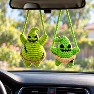 Puede incluir: Dos figuras de ganchillo de color verde lima colgadas del espejo retrovisor de un coche. Cada figura tiene una cabeza puntiaguda, ojos cosidos en negro y una boca ancha y dentada. La figura más grande tiene detalles cosidos en negro y una faja beige.