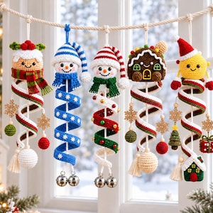 Op de afbeelding: Vijf hangende gehaakte kerstornamenten met een pudding, twee sneeuwpoppen, een peperkoekhuisje en een geel figuurtje. Elk ornament heeft een spiraalvormige staart met houten sneeuwvlokken, kleine gekleurde bolletjes en metalen belletjes.