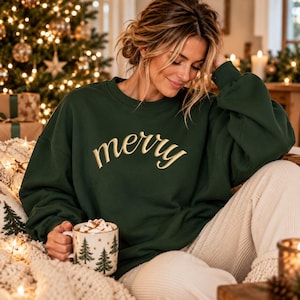 May include: A dark forest green crewneck sweatshirt featuring the word merry embroidered in a gold cursive font across the chest. The garment has long sleeves and a relaxed fit, providing a comfortable look for the holiday season.