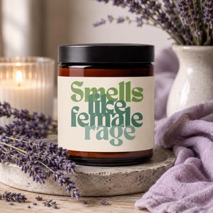 May include: A brown glass candle jar with a black lid and a cream-colored label. The label features the text Smells like female rage in stacked, wavy green and teal typography. The candle is positioned among dried lavender sprigs.