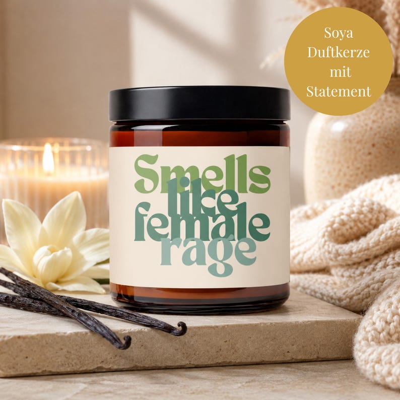 May include: A brown glass candle jar with a black lid and a cream-colored label featuring the green text "Smells like female rage" in a bold, wavy font. The candle is displayed alongside a white flower and two dark vanilla bean pods.