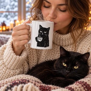 May include: A white ceramic mug featuring a black ink illustration of a grumpy cat holding a small cup with the text "Stuff" printed on it. The mug has a standard handle and a smooth finish, showcasing a detailed graphic of a feline.