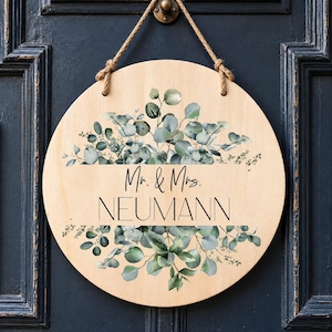 May include: A round light-colored wooden sign hanging by a jute rope. The sign features a printed design of green eucalyptus leaves and branches. In the center, black text reads "Mr. & Mrs." above the name "NEUMANN" in a larger font.