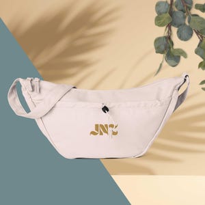 May include: A light beige crescent-shaped crossbody bag featuring a smooth fabric exterior, a wide shoulder strap, and two top zipper closures. The front includes a drawstring pocket with a black toggle and the gold embroidered text JNC.