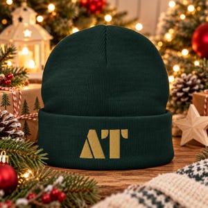 May include: A dark forest green knit beanie featuring a folded cuff. Embroidered in the center of the cuff is the gold-colored text AT in a bold, sans-serif font. The hat is constructed from a ribbed fabric with a classic rounded crown.