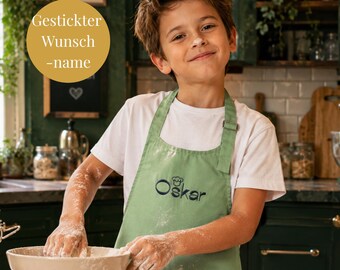 Può includere: Un grembiule verde salvia con il nome Oskar ricamato in filo blu scuro, accompagnato da una piccola icona di un cappello da chef sopra il testo. Il grembiule è indossato sopra una t-shirt bianca semplice, con un cerchio dorato sovrapposto contenente il testo Gestickter Wunsch-name.