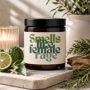 May include: A dark amber glass candle jar with a black lid and a cream-colored label. The label features the green text "Smells like female rage" in a bold, wavy font. The candle is positioned next to a sliced lime and green foliage.