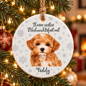 May include: A round white ceramic ornament featuring a brown and white puppy illustration, gray snowflake patterns, and the black text "Unser erstes Weihnachtsfest mit" above the puppy and the name "Teddy" printed below it.