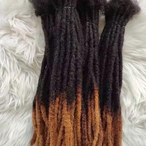 May include: A bundle of synthetic dreadlocks featuring a two-tone color gradient. The roots are a deep, dark black that transitions into a warm, light brown shade toward the ends. The texture is coiled and dense, with individual strands bundled together.