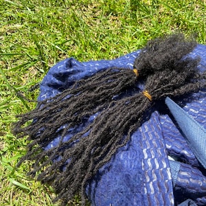 May include: Two bundles of dark black hair extensions styled into thin, textured dreadlocks. Each bundle is secured at the top with a small, thin gold-colored elastic band. The hair strands are long, thin, and have a coarse, matted appearance.