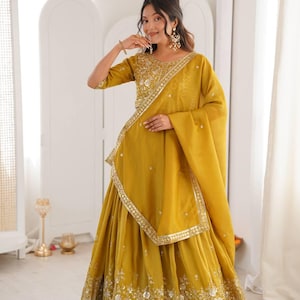 May include: Mustard yellow traditional Indian outfit. The ensemble includes a top with intricate gold embroidery, a matching skirt, and a draped scarf. The skirt and top feature detailed silver embellishments. The outfit is worn by a person.