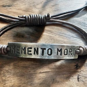 Puede incluir: Una pulsera de color plateado con las palabras "MEMENTO MORI" grabadas en una placa rectangular. La pulsera está hecha de un cordón de cuero marrón, anudado en los extremos, y se muestra sobre una superficie de madera.