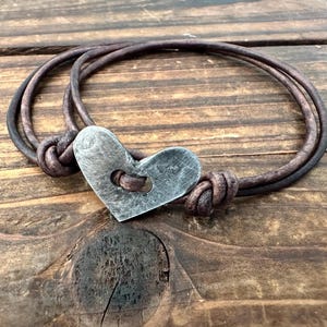 Puede incluir: Una pulsera de cuero marrón con un cierre metálico en forma de corazón. La pulsera está hecha de tres hebras de cuero y está anudada a cada lado del corazón. El cierre de corazón tiene un agujero en el centro.