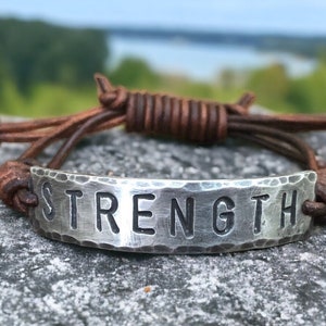 May include: A silver bracelet with the word "STRENGTH" stamped on a hammered metal plate. The bracelet features a brown leather cord that wraps around the wrist. The background is a blurred view of nature.