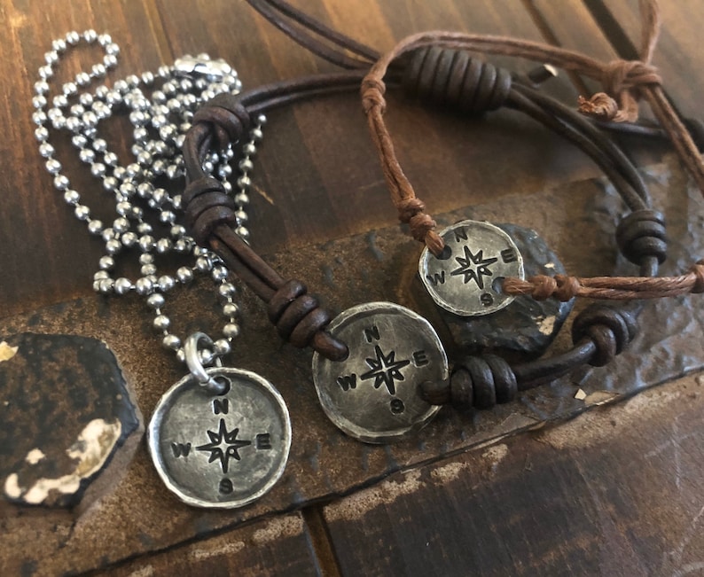 Puede incluir: Dos collares de cord&oacute;n de cuero con colgantes de br&uacute;jula. Los colgantes son de plata y presentan una rosa de los vientos con las letras N, S, E y W.