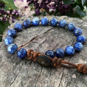 K&ouml;nnte beinhalten: Ein blaues Lapislazuli-Perlenarmband mit einem braunen Lederband und einem Holz-Knopfverschluss.
