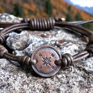 Puede incluir: Un brazalete de cuero marr&oacute;n con un dije de br&uacute;jula. La br&uacute;jula es de metal y tiene un dise&ntilde;o de estrella en el centro. El brazalete est&aacute; atado con un nudo.