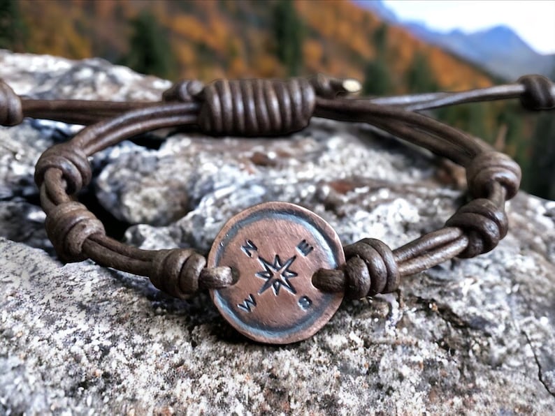 Puede incluir: Un brazalete de cuero marr&oacute;n con un dije de br&uacute;jula. La br&uacute;jula es de metal y tiene un dise&ntilde;o de estrella en el centro. El brazalete est&aacute; atado con un nudo.