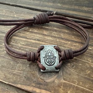 Könnte beinhalten: Ein braunes Lederarmband mit einem rechteckigen Silberanhänger mit einem Hamsa-Hand-Design. Das Armband hat mehrere Lederstränge und ist zur Anpassung geknotet. Der Anhänger ist mittig auf dem Armband angebracht.