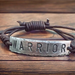 May include: A brown leather bracelet with a silver metal tag that says "WARRIOR".