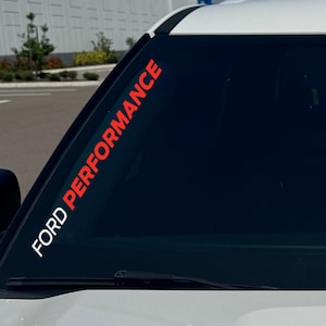 Op de afbeelding: Een witte en rode vinyl sticker aangebracht op het bovenste gedeelte van de voorruit van een voertuig. De sticker bevat de tekst FORD PERFORMANCE in een vet, schreefloos lettertype, waarbij FORD in het wit en PERFORMANCE in het rood is.