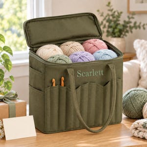 May include: An olive green rectangular yarn storage bag with a zipped lid, featuring the name Scarlett embroidered on the front. The bag has multiple exterior pockets containing two wooden crochet hooks and is filled with six balls of yarn.