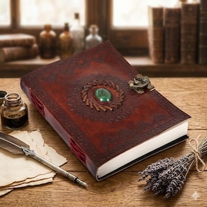 Op de afbeelding: Een bruin leren dagboek met een reliëfontwerp en een groene stenen middelpunt. Het dagboek heeft een metalen sluiting en rode binding. Een ganzenveer, een inktfles en gedroogde lavendel zijn ook zichtbaar.