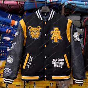 Könnte beinhalten: Schwarze und goldene Collegejacke mit einem Bulldoggen-Patch und den Buchstaben "AT". Die Jacke hat Lederärmel und den Text "EST.1891" und "Got Love For My HBCU".