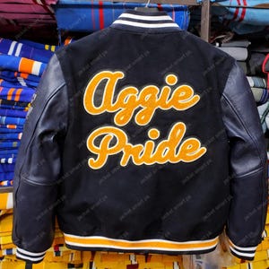 Puede incluir: Chaqueta universitaria negra con mangas de cuero. La espalda presenta las palabras "Aggie Pride" en letras naranjas y blancas. La chaqueta tiene ribetes a rayas blancas y naranjas en los pu&ntilde;os y el dobladillo.