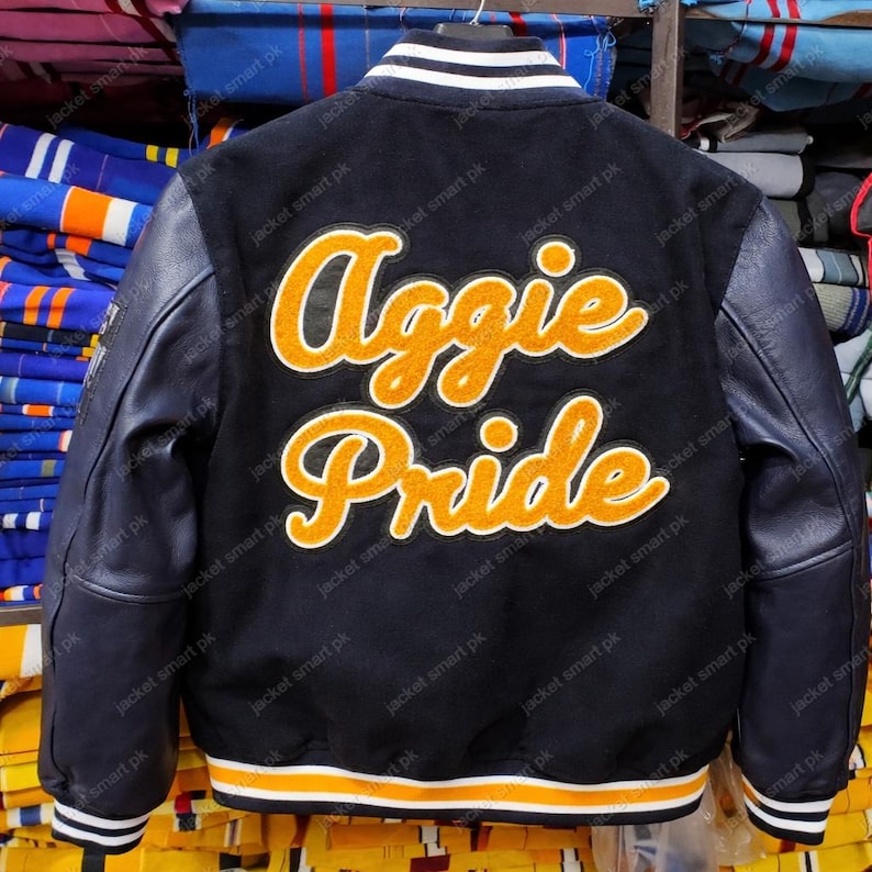 Puede incluir: Chaqueta universitaria negra con mangas de cuero. La espalda presenta las palabras "Aggie Pride" en letras naranjas y blancas. La chaqueta tiene ribetes a rayas blancas y naranjas en los pu&ntilde;os y el dobladillo.