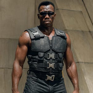 May include: A man wearing a black tactical vest with multiple pockets and silver buckles, black sunglasses, and black trousers. The vest has a zip-up front and is worn over a bare chest. The background is a light-coloured concrete structure.