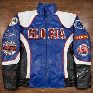 Op de afbeelding: Een blauw, wit en zwart leren jack met het woord "GLORIA" in rode blokletters over de borst. De jas heeft meerdere patches en het jaartal "2025" op de mouwen. De jas heeft een ritssluiting en een opstaande kraag.