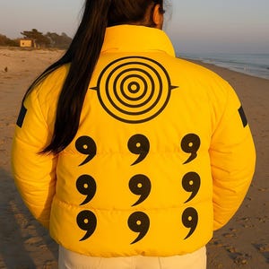 Puede incluir: Chaqueta acolchada amarilla con gráficos negros. La chaqueta presenta un gran diseño circular en la parte superior de la espalda y nueve símbolos del número nueve dispuestos en un patrón de cuadrícula en la parte inferior de la espalda.