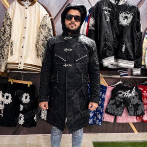 May include: A black hooded jacket with a quilted design on the shoulders and upper chest. The jacket features multiple silver zippers and clasps, and is worn with blue jeans. Other jackets and shorts are visible in the background.