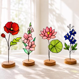 以下が含まれることがあります： 丸い木製の台座に乗った4つのステンドグラス製の花の彫刻。左から順に、緑の葉を持つ赤いポピー、ピンクのグラジオラス、緑のハスの葉を持つピンクの蓮の花、そして緑の葉を持つ青いデルフィニウムが並んでいます。